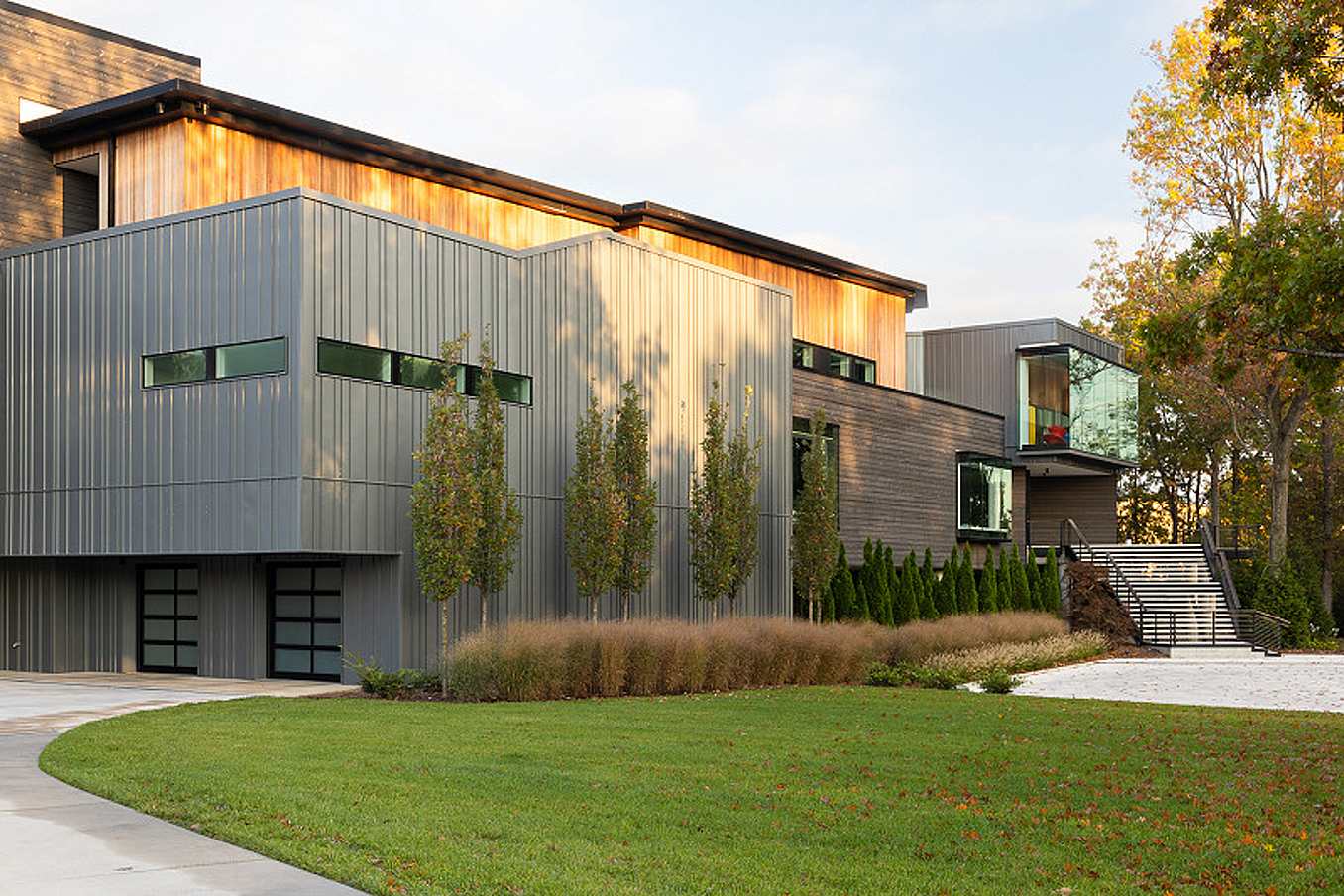 Modern two-story home with metal and wood exterior, large transom windows, and landscaped front yard, daytime driveway view.