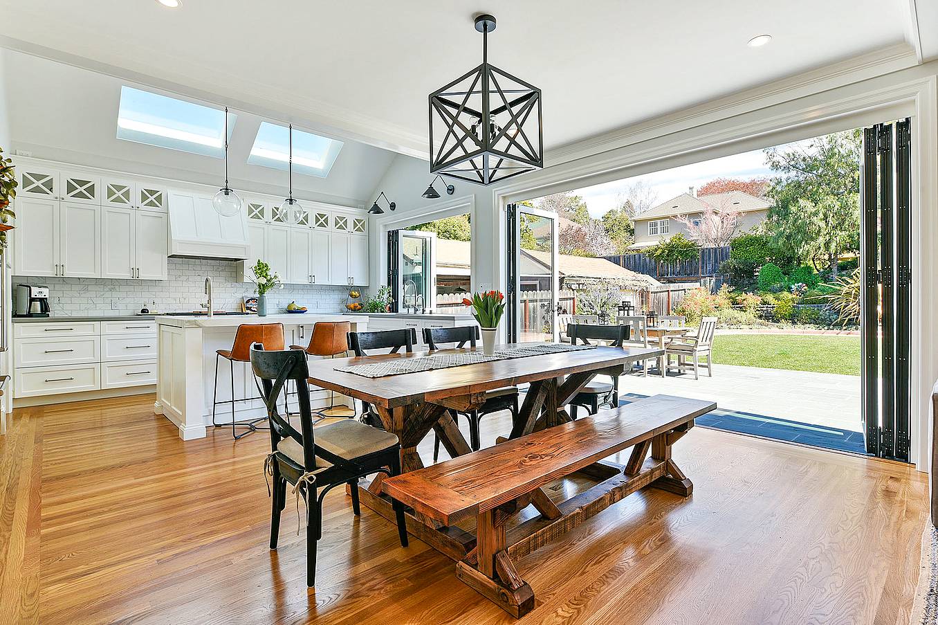 Contemporary open-plan kitchen and dining area with wooden floors, modern white cabinets, skylights, and folding glass walls leading to a backyard patio.