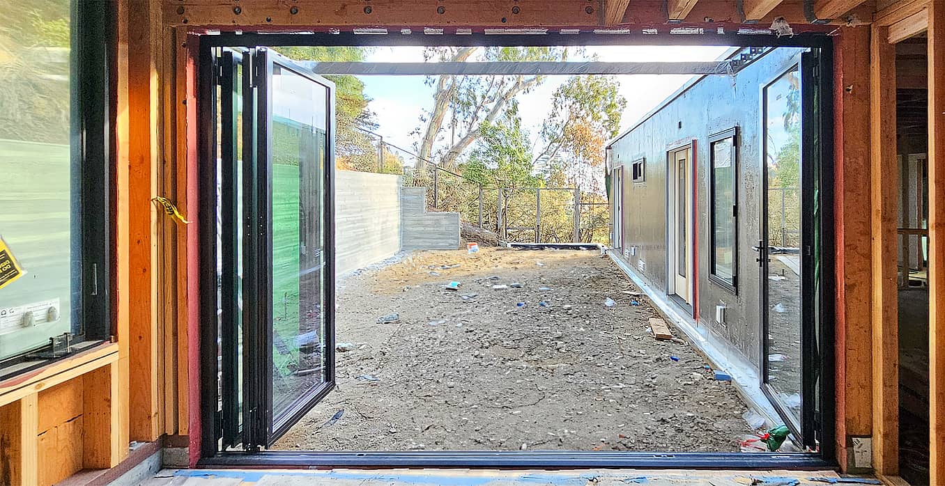 Open aluminum clad folding glass wall reveals a yard under construction, framed by wooden walls and trees in the background.