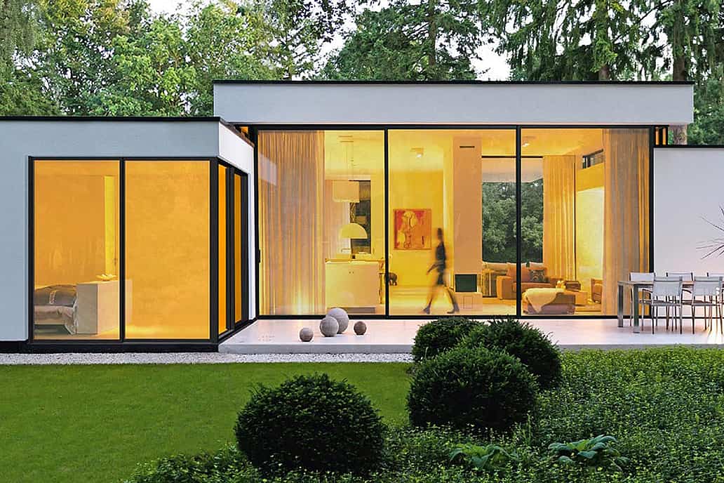A modern house features large glass windows and a sleek cero minimal sliding glass door system, glowing warmly from indoor lights.