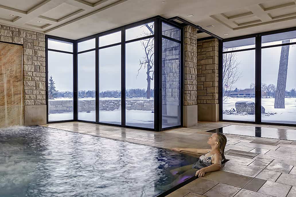 Modern indoor pool with stone waterfall and glass doors overlooking snowy landscape