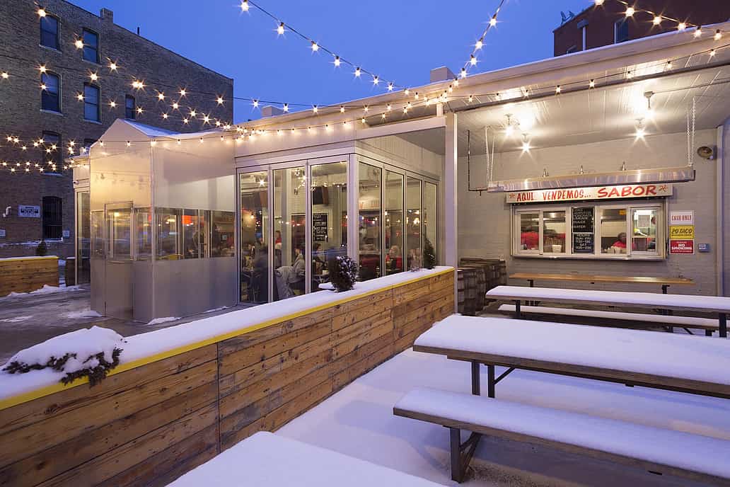 Outdoor restaurant patio with string lights, snow-covered seating, and high-performance glass doors showcasing a cozy indoor restaurant space.