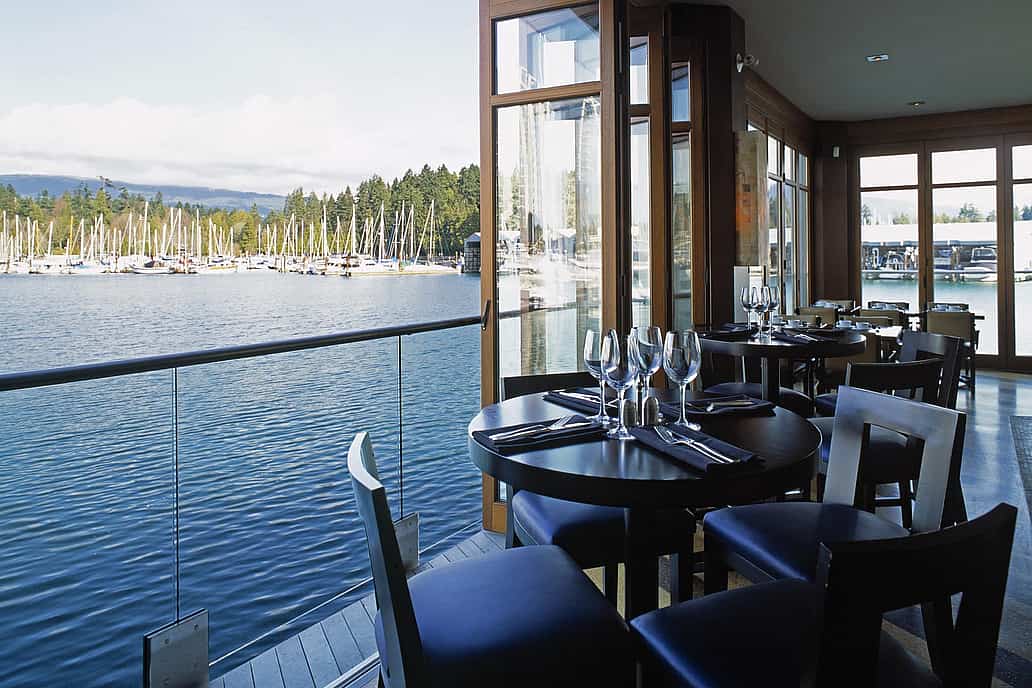 Indoor restaurant seating with set tables and waterfront views of a marina with sailboats, seen through expansive sliding glass doors.
