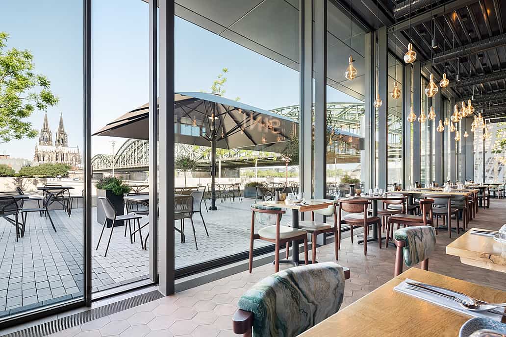 Modern restaurant with floor-to-ceiling glass doors, cityscape views of a cathedral and bridge, and patio seating.