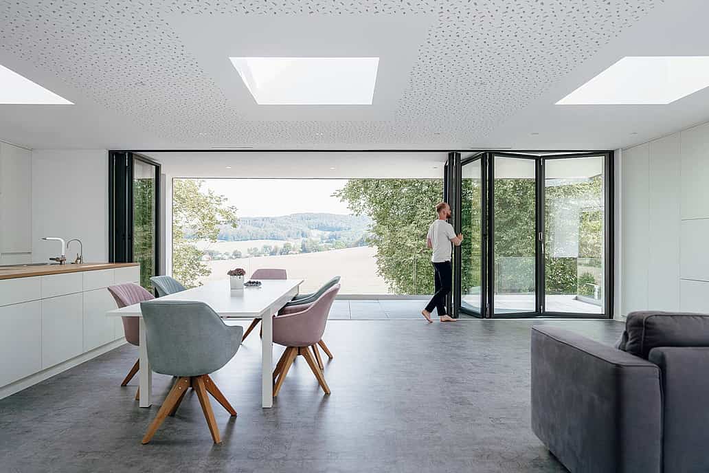 A person opens folding glass doors in a modern, open-plan living and dining area, seamlessly connecting the space to scenic views of trees and fields outside.