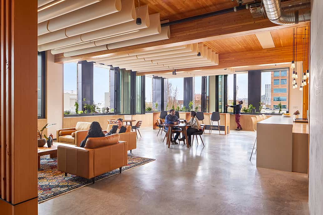 Modern office lounge with sofas, a table, wooden ceiling, indoor plants, and large folding windows for ample natural light.