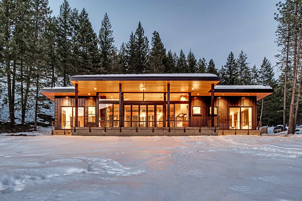 Modern cabin with large windows and warm interior lighting, surrounded by snow and tall pine trees.