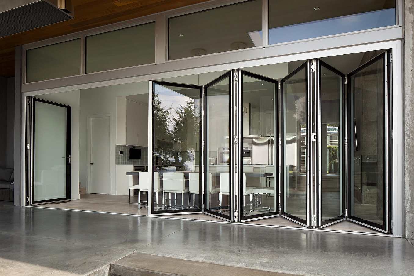 Glass folding doors open to reveal a modern kitchen and dining area. White cabinets and a table with chairs are visible through the doors.