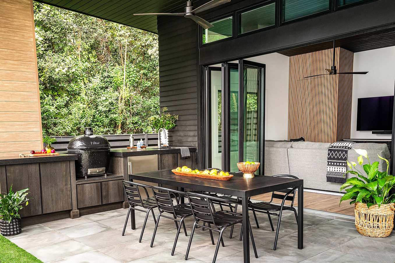 Modern outdoor patio with black dining table and six chairs, built-in grill, countertop, and and large folding glass doors opening to a living room with a sofa and TV.