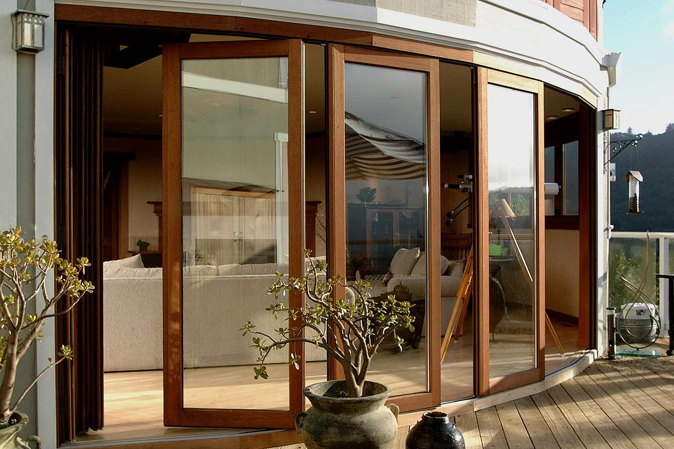 Curved wood-framed sliding glass doors open from a modern living room to a patio with potted plants and scenic views of trees and rolling hills.