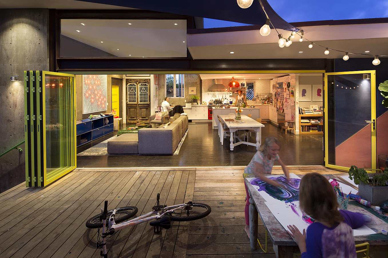 Two children make art on a wooden deck near accordion glass doors opening to a modern, well-lit living and kitchen area.