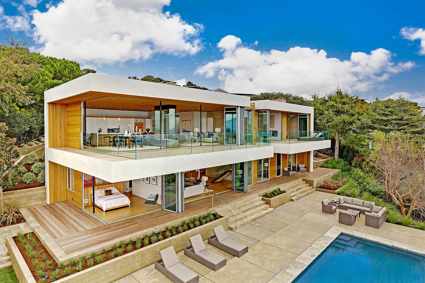 Modern two-story house with large sliding glass doors, open balconies, poolside loungers, and outdoor seating in landscaped surroundings.