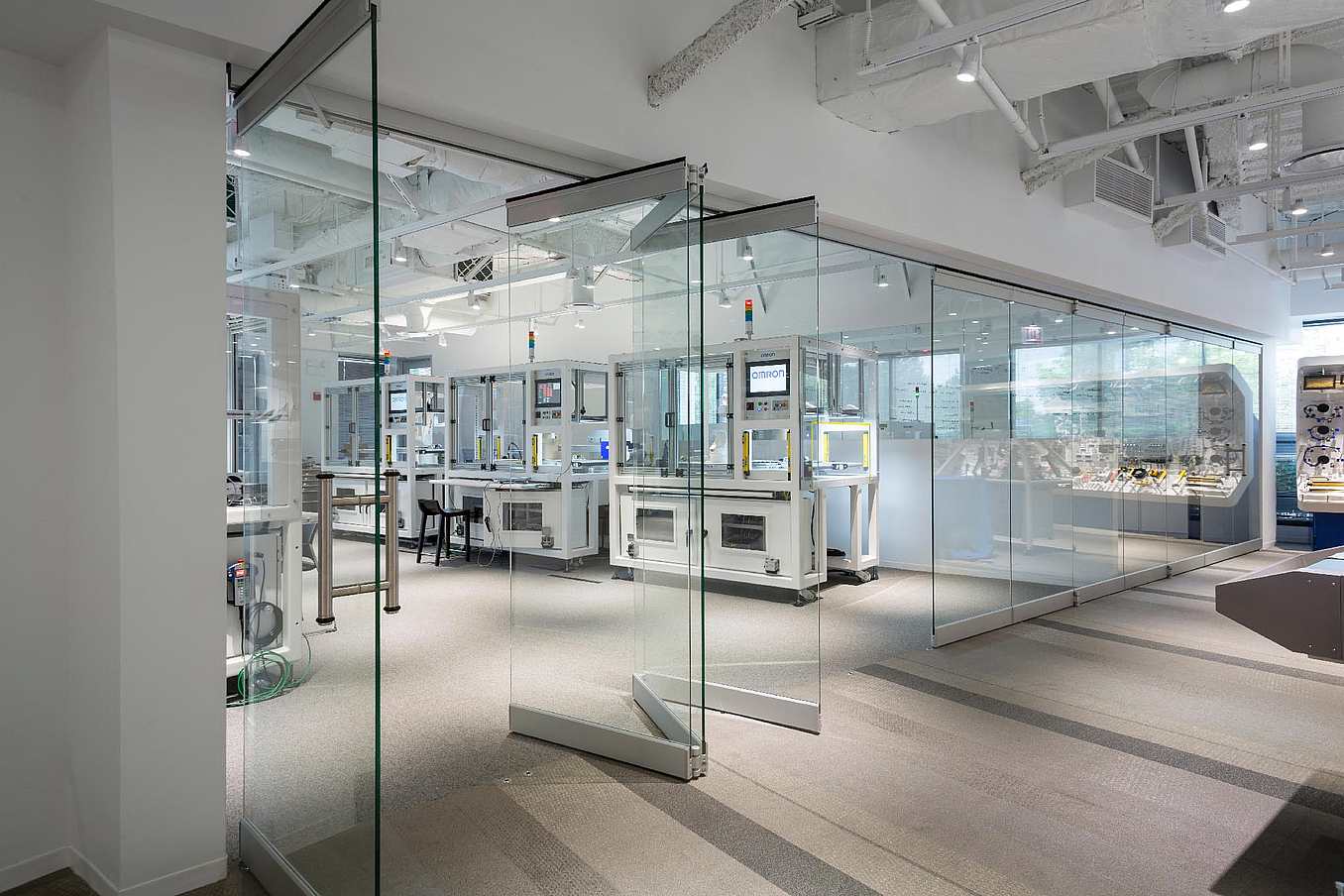 A modern laboratory with frameless folding glass doors, glass walls, workstations, computers, and scientific equipment, well-lit by ceiling lights and natural light from windows.