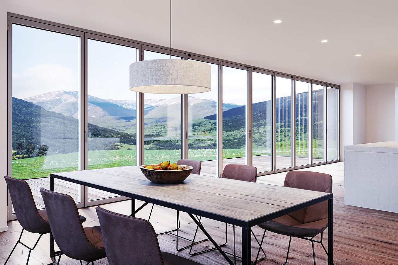 Modern dining room and kitchen with a wooden table, six chairs, and a fruit bowl, featuring floor-to-ceiling glass doors overlooking a mountain landscape.