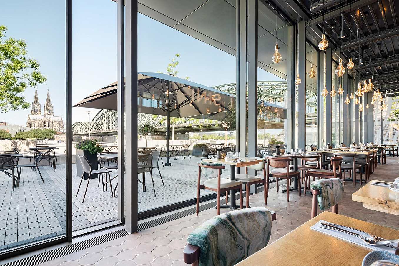 Modern restaurant with floor-to-ceiling glass doors, cityscape views of a cathedral and bridge, and patio seating.