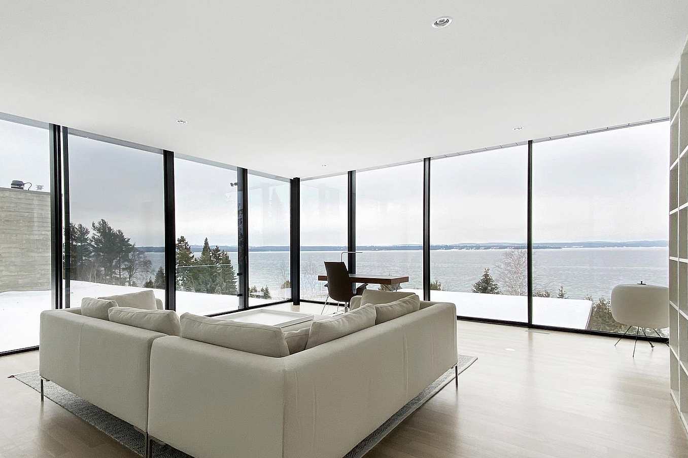 Minimalist living room with a white sofa, sleek floor-to-Ceiling sliding glass doors, serene snowy and water views, light wood flooring, and modern built-in shelving.