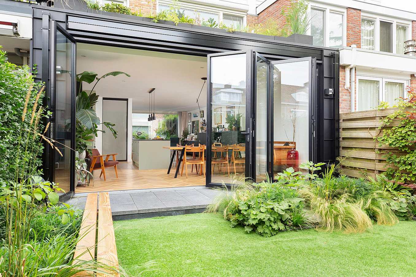 Modern home with large glass doors opening to a green garden, showing a spacious, light-filled kitchen and dining area with wooden flooring and contemporary furniture.
