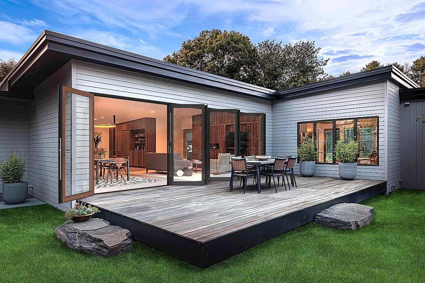 Modern house with large aluminum clad bifold glass doors opens to a wooden deck featuring an outdoor dining set, surrounded by green grass and potted plants.