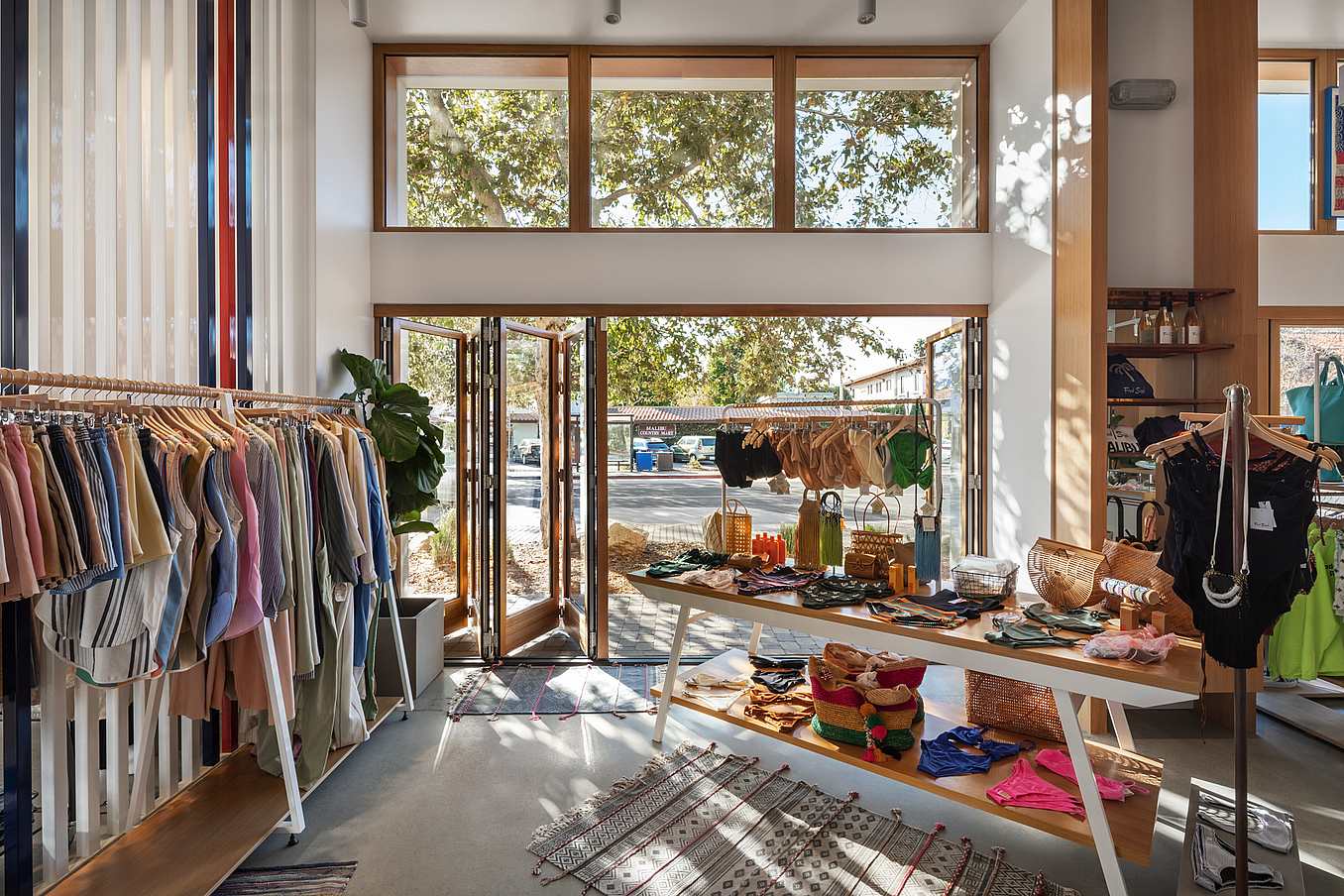 A brightly retail store features clothes on racks and accessories on tables, enhanced by accordion glass doors that offer a seamless view of trees through large windows and an open door.