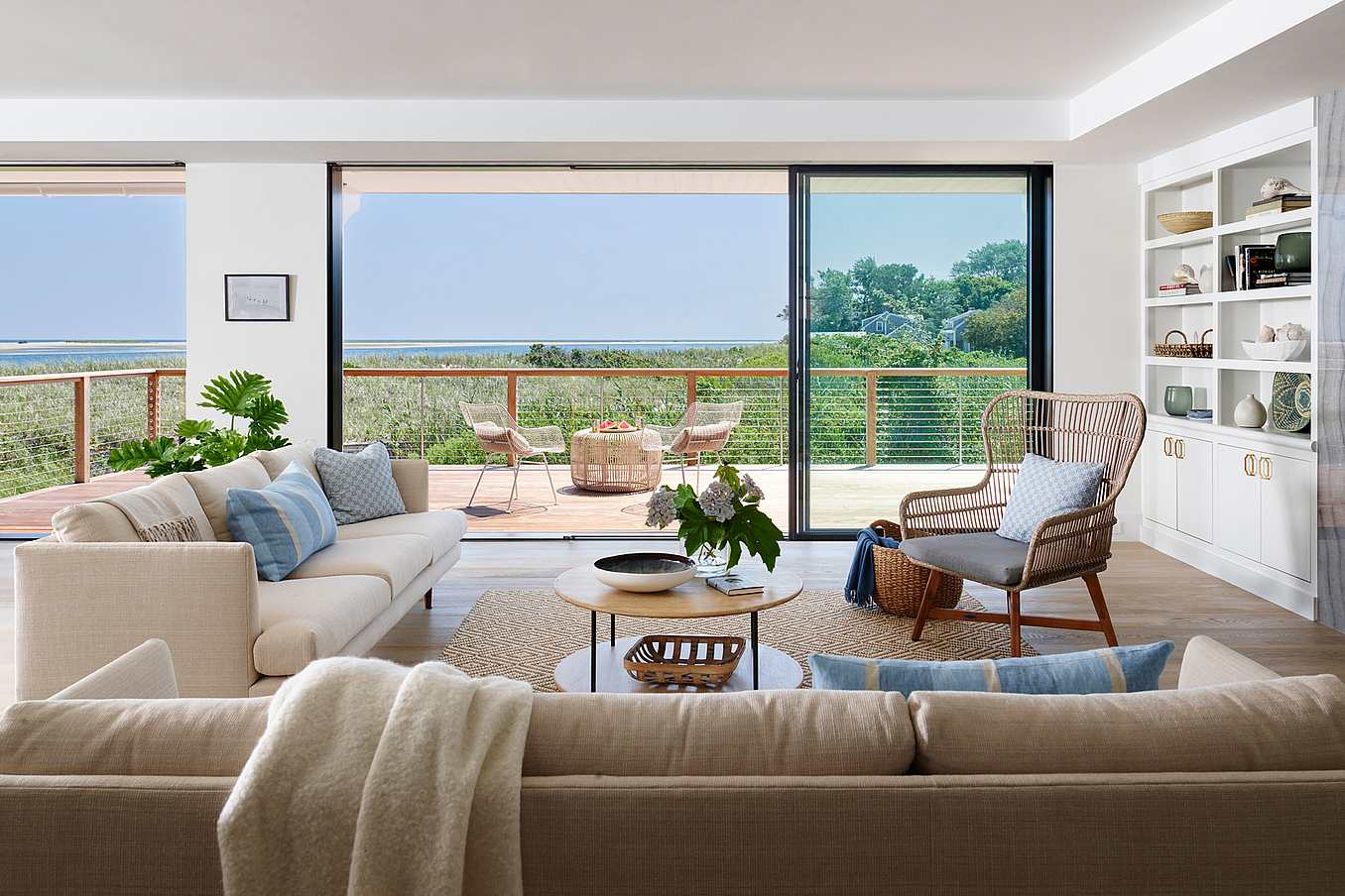 Modern living room with neutral furniture, light wood accents, and built-in shelves, featuring sliding glass doors opening to a deck with outdoor seating.