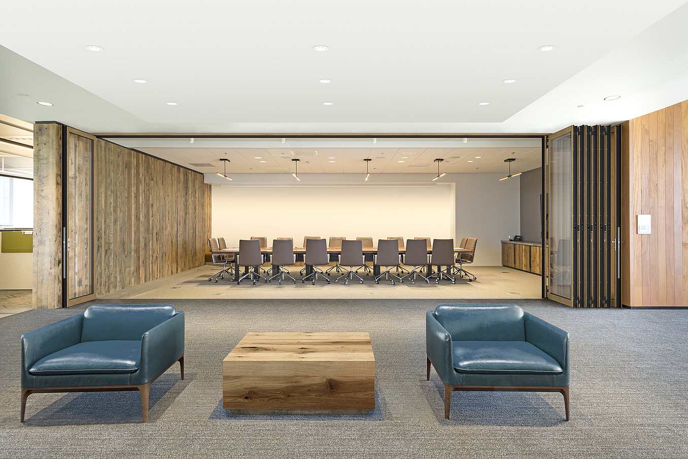 Modern office lobby with two blue armchairs and a wooden coffee table, facing a conference room opened folding glass doors.