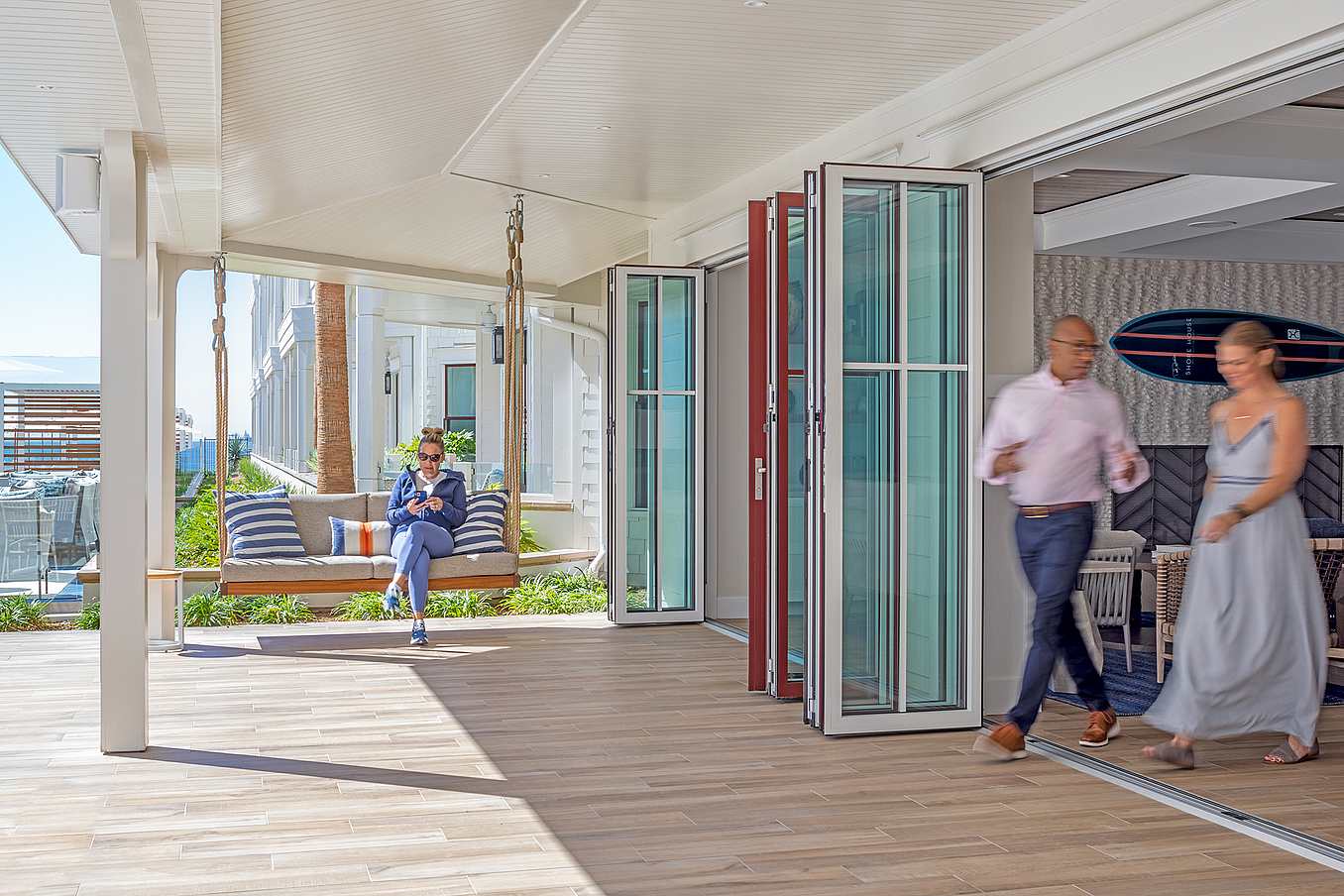 A man sits on a suspended outdoor bench while a man and a woman walk through an open folding glass door into a bright, modern hotel lobby area.