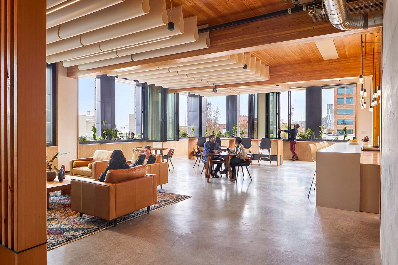 Modern office lounge with sofas, a table, wooden ceiling, indoor plants, and large folding windows for ample natural light.