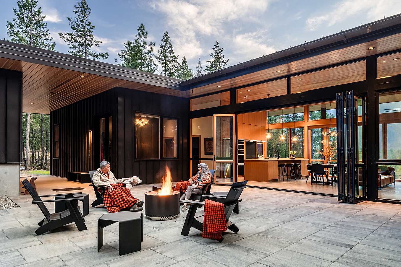 Two people sit by a fire pit on a modern patio, surrounded by tall trees. The house features large accordion glass doors and an open living area visible in the background.