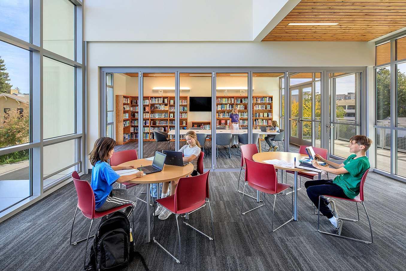 Modern library interior with opening glass doors, large windows, round tables with red chairs, and bookshelves.