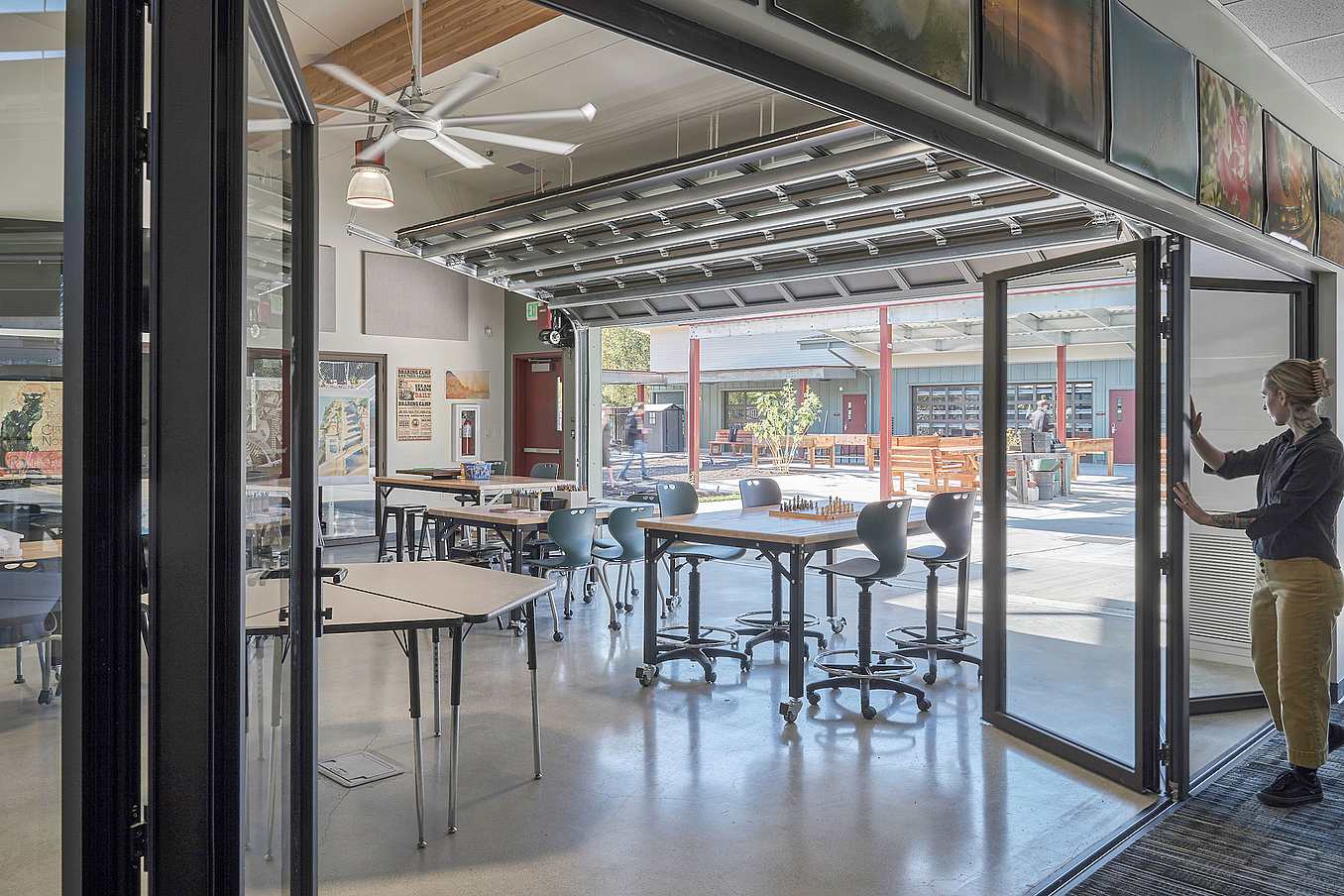 A person slides open a glass partition in a bright classroom with tables, chairs, and an open-air area with additional seating.