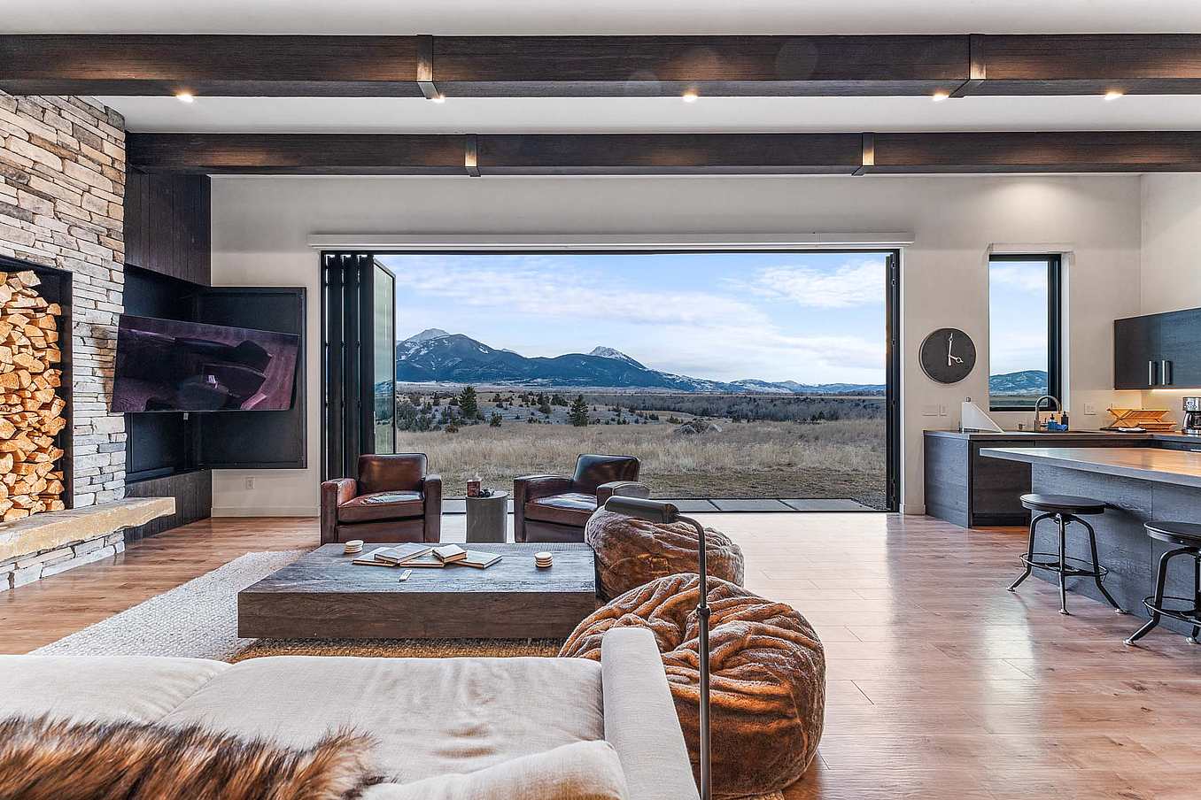 Bifold glass doors open a modern living room with stone fireplace, wood accents, and kitchen bar, overlooking mountain views