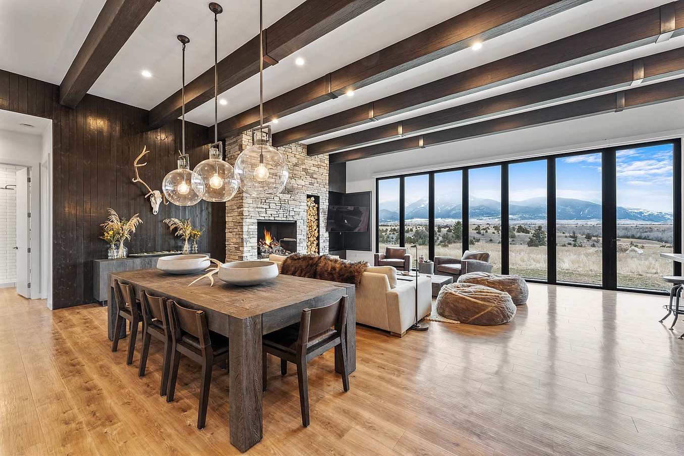 Bifold glass doors frame a modern living and dining area with wood floors, stone fireplace, and floor‑to‑ceiling windows overlooking scenic mountains