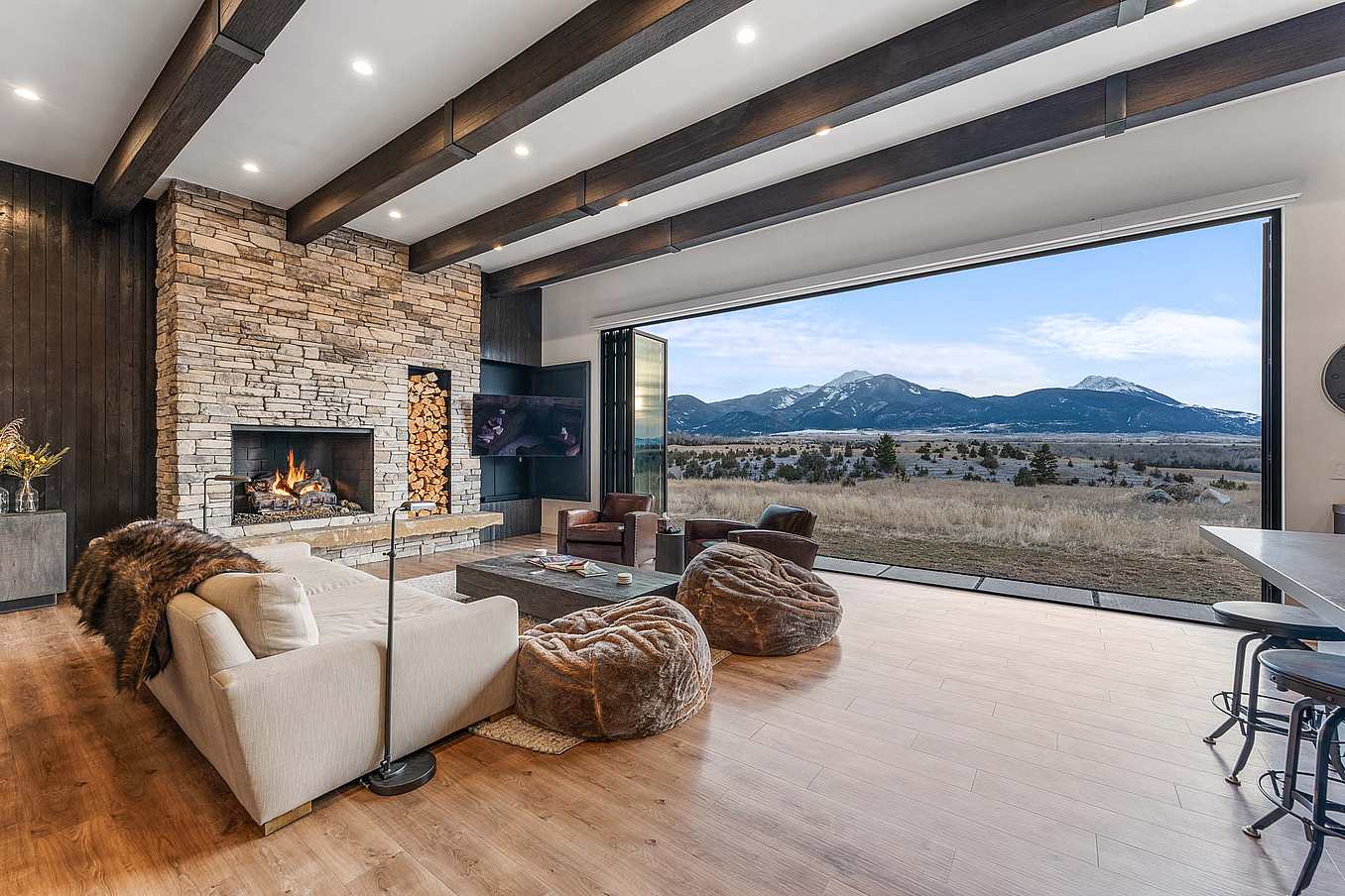 Bifold glass doors connect a modern living room with stone fireplace and wood floors to scenic mountain views, creating seamless indoor-outdoor living