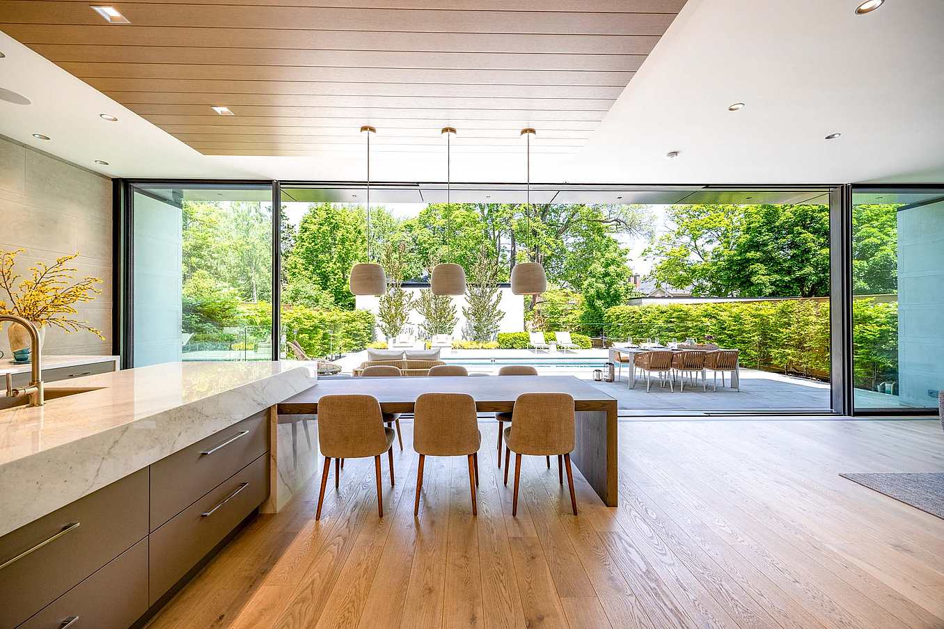 Large sliding glass doors connect a modern kitchen and dining area to a backyard patio with pool, outdoor dining, and lush greenery