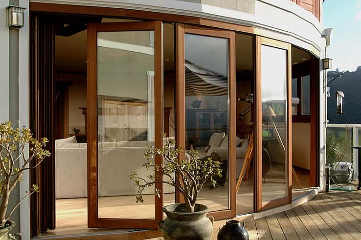 Curved wood-framed sliding glass doors open from a modern living room to a patio with potted plants and scenic views of trees and rolling hills.