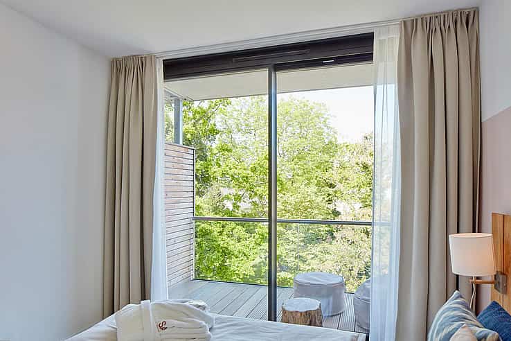 Modern bedroom with double bed, bedside lamp, robes, and floor-to-ceiling windows opening to a balcony with stools and tree views.