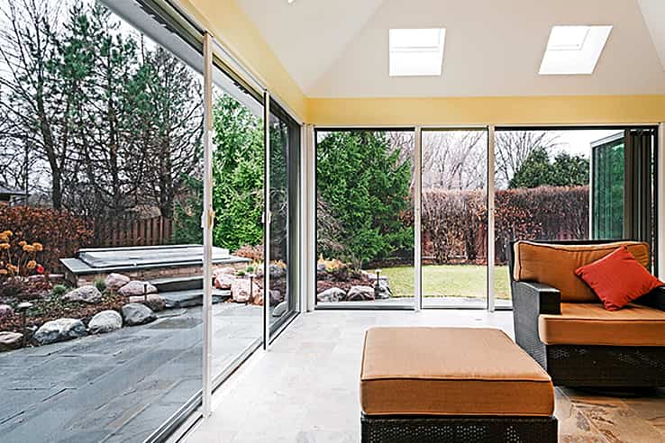 Sunroom with sliding glass doors and screens opens to a landscaped backyard with trees.