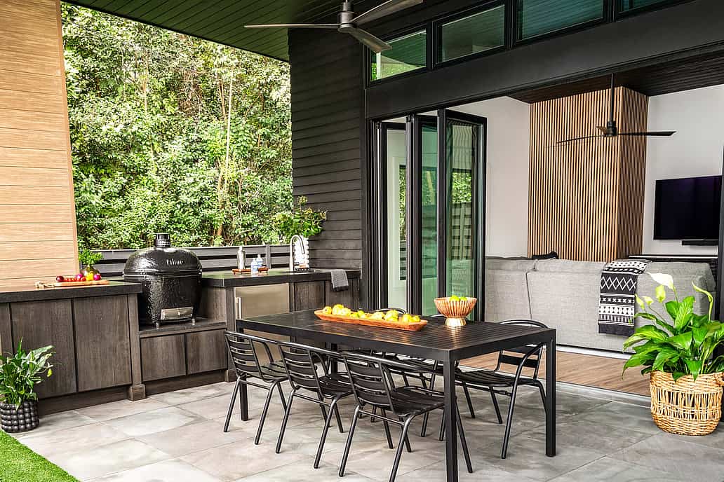Modern outdoor patio with black dining table and six chairs, built-in grill, countertop, and and large folding glass doors opening to a living room with a sofa and TV.