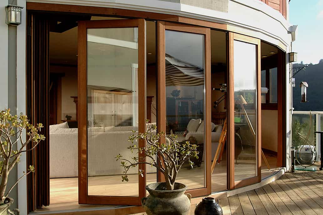 Curved wood-framed sliding glass doors open from a modern living room to a patio with potted plants and scenic views of trees and rolling hills.