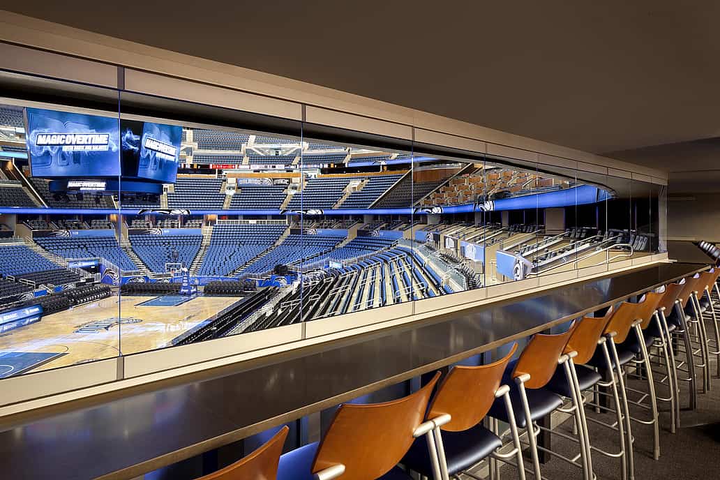 Orlando Magic suite with seating and frameless sliding glass windows, overlooking the indoor basketball arena and scoreboard.