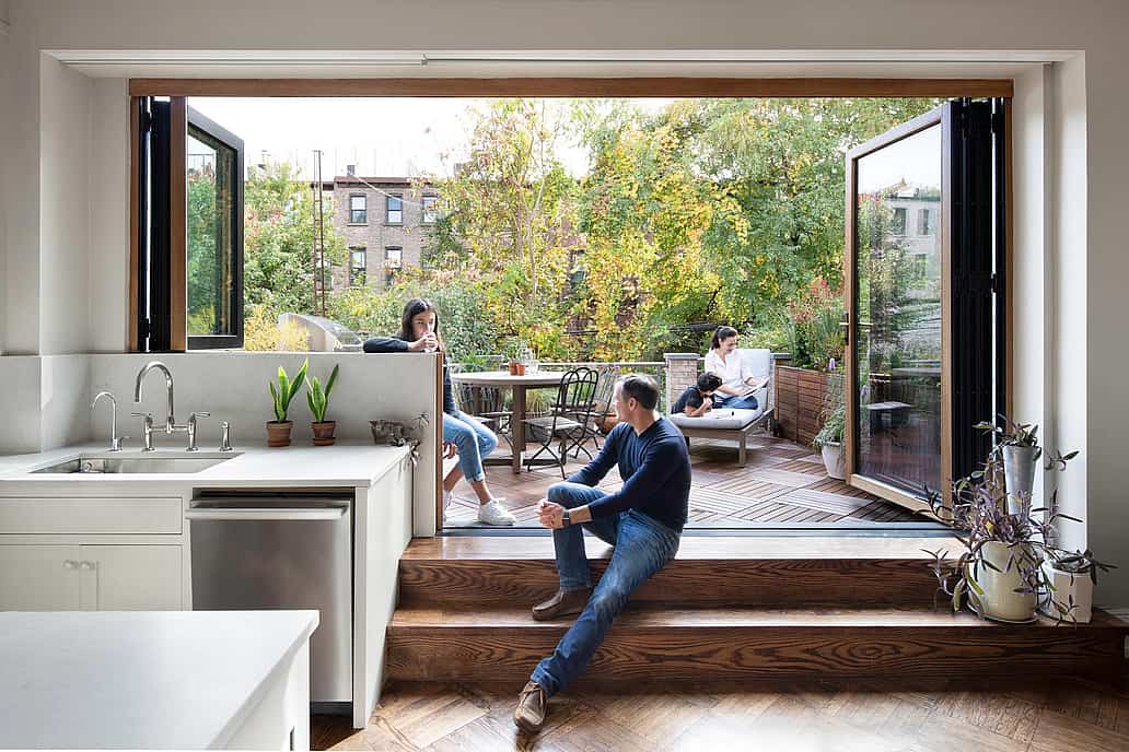 Modern kitchen with retractable glass wall that opens the kitchen to a patio. Three people relax in a modern home with a retractable glass wall that opens the kitchen to a patio; two people sit outside amid greenery, while one sits on indoor steps next to houseplants.