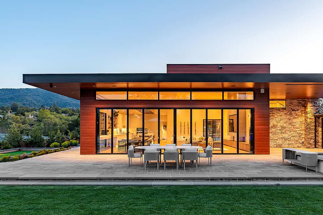 Modern home with transoms windows and floor-to-ceiling folding patio doors, featuring stylish outdoor furniture and sunset views over lush hillside scenery.