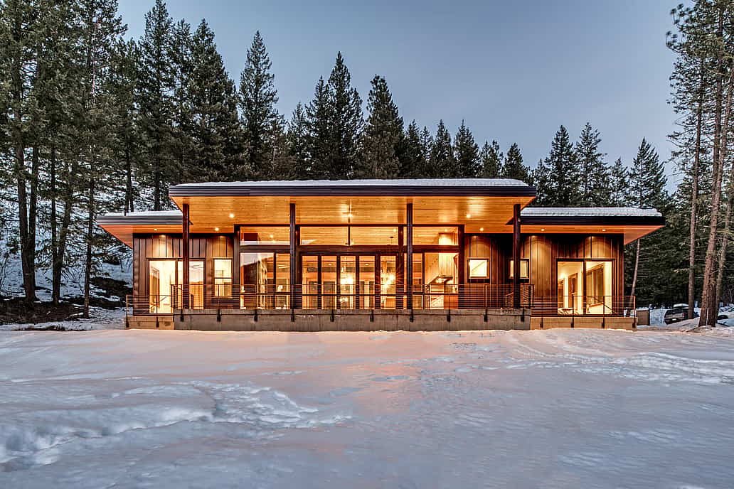 Modern cabin with large windows and warm interior lighting, surrounded by snow and tall pine trees.