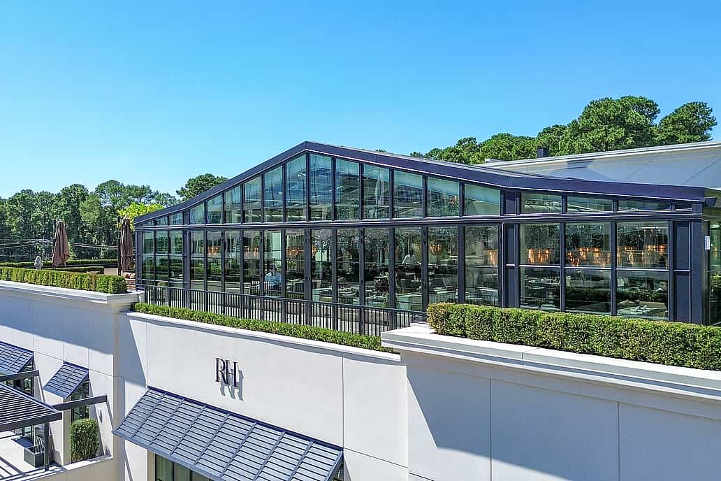 Restoration Hardware rooftop restaurant in Raleigh NC featuring NanaWall SL70 bifold glass doors as the focal point of a modern glass-enclosed dining space with a greenhouse-style roof surrounded by greenery under a clear blue sky