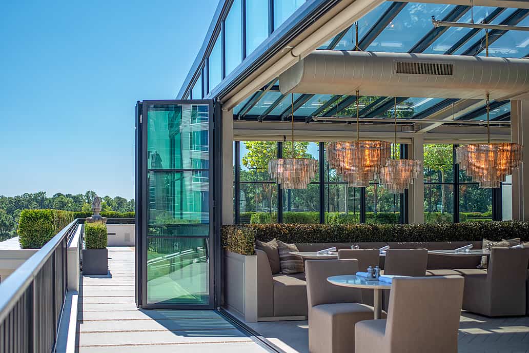 Restoration Hardware rooftop restaurant in Raleigh NC featuring NanaWall SL70 bifold glass doors, glass walls and ceiling, outdoor seating with chandeliers, and views of greenery under a clear blue sky