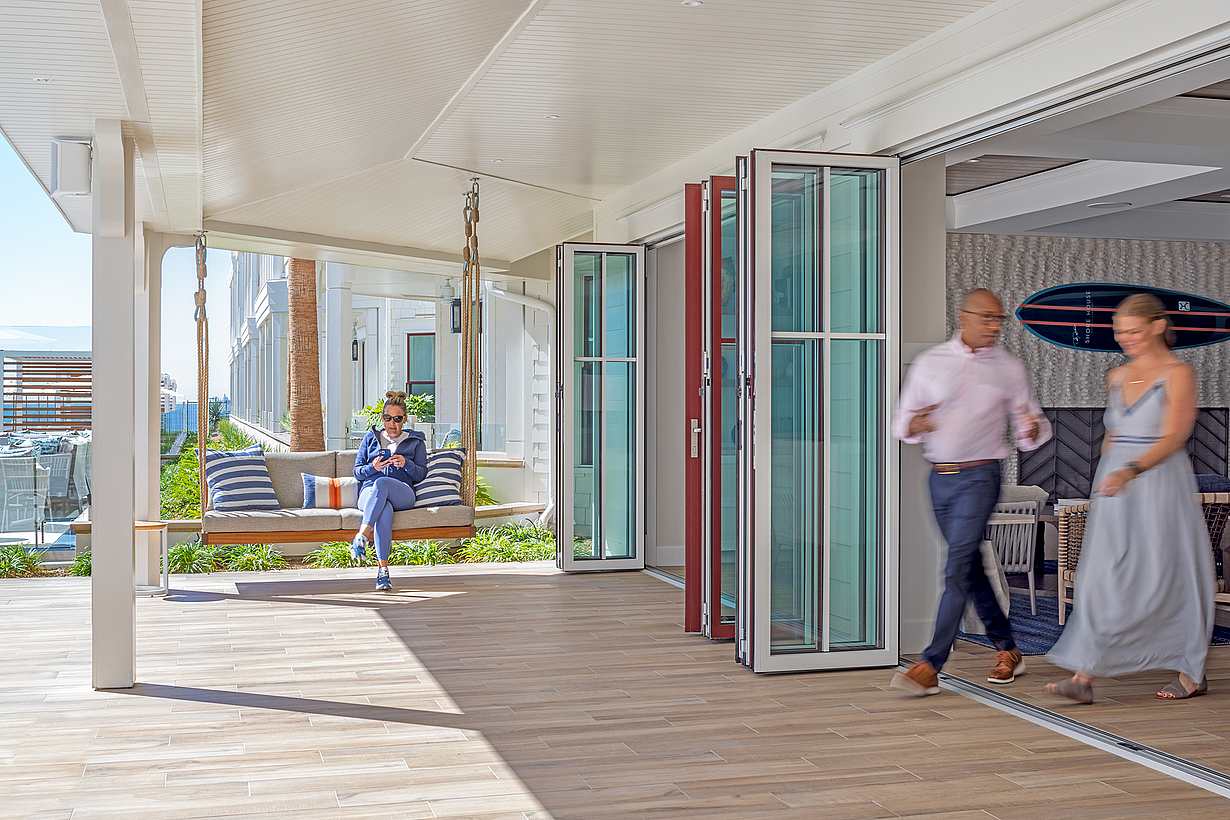 A man sits on a suspended outdoor bench while a man and a woman walk through an open folding glass door into a bright, modern hotel lobby area.