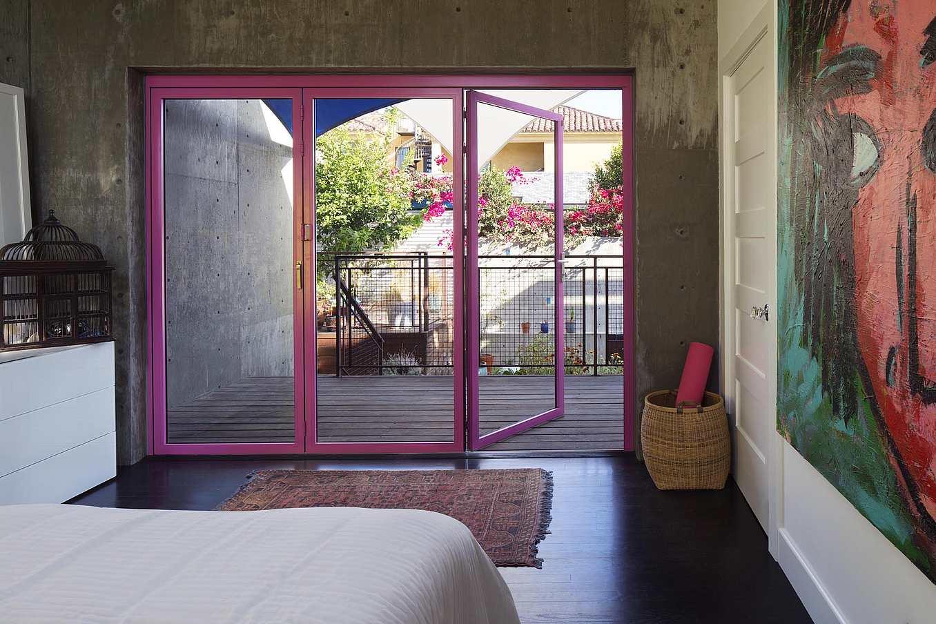 Modern bedroom with concrete accent wall, custom-colored glass swing doors opening to a balcony, and a vibrant abstract painting on the side wall.