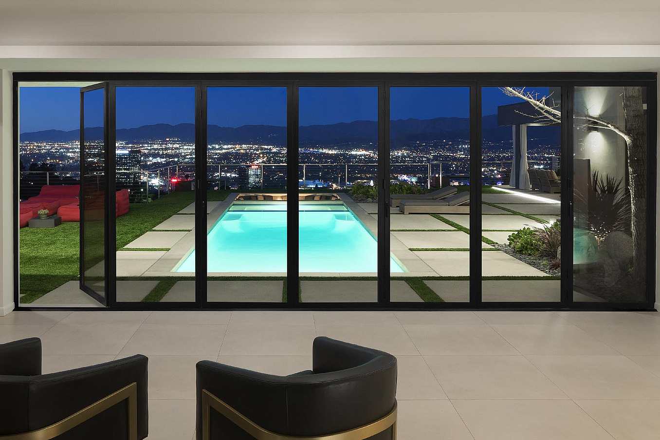 Modern living room interior with swing glass doors opening to a luxury pool, stylish patio, and panoramic nighttime cityscape view with mountain backdrop.