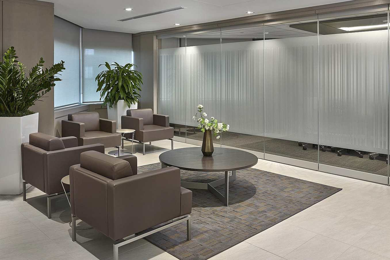 Modern office waiting area with brown armchairs and a round coffee table, leading to a conference room with frosted frameless sliding glass doors.