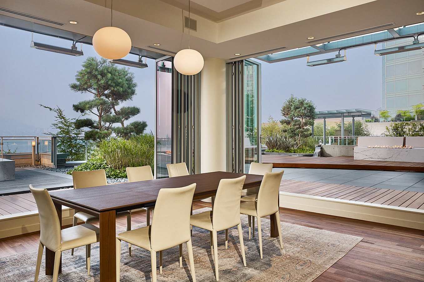 Modern dining area with a wooden table and eight white chairs, featuring accordion glass doors that open to a spacious outdoor patio with greenery.